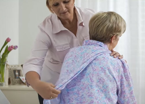 Middle aged female carer is helping an elderly woman with a floral-patterned petal back nightie in a care home setting.