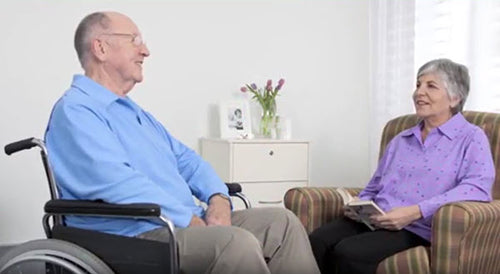 Photo of an older man in a wheelchair, wearing a light blue petal back long-sleeved polo shirt. He is sitting opposite an older woman in a chair. She is wearing a petal back long-sleeved tunic blouse in a purple pattern.